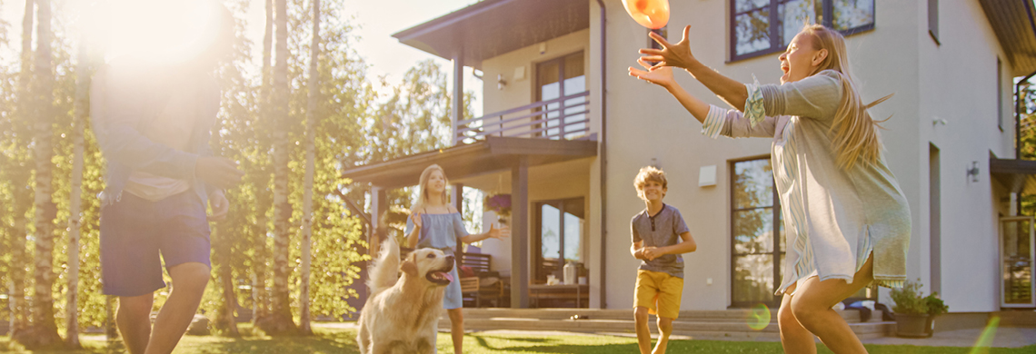 Familie spielt mit einem Ball und einem Hund im Garten vor einem modernen Haus bei Sonnenuntergang.