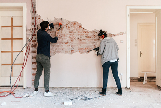 Zwei Personen entfernen Putz von einer Wand, um die darunter liegenden Ziegel freizulegen; der Boden ist mit Schutt bedeckt.
